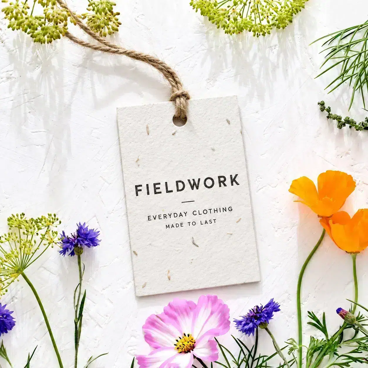Seed paper swing tag with jute string, printed with a clothing brand logo, surrounded by wildflowers